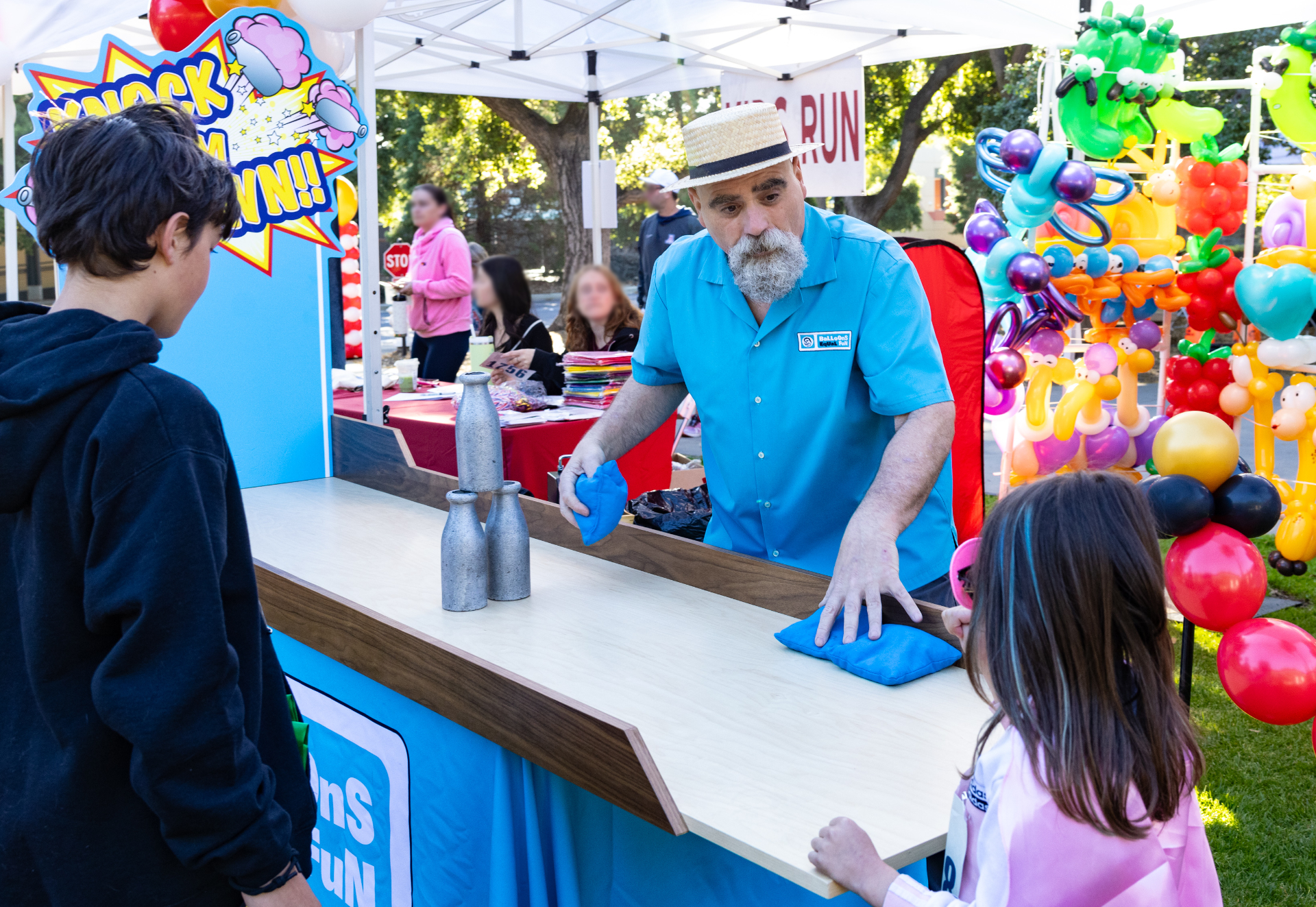 The carnival game 'Knock 'Em Down!' There are 3 classic milk cans, stacked on a wooden tabletop alley. Brian is explaining the gameplay while handing a beanbag to a young player while her brother looks on.